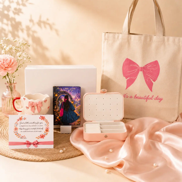 A styled gift set featuring a beige canvas tote bag with a pink bow graphic and 'it's a beautiful day' text, a blush pink jewelry organizer case, a white gift box, a small card, a mug, and floral decor on a soft beige background.