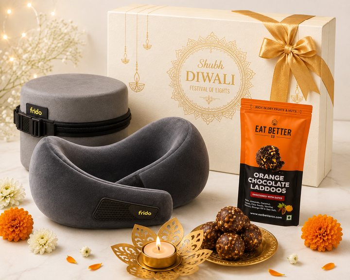 A Frido travel neck pillow set displayed with its branded gift box, alongside an orange bag of Eat Better Chocolate Laddos, a small lit candle, and decorative flowers on a light neutral background.