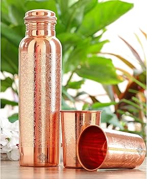 A copper-colored metal tumbler with a textured pattern, shown alongside two matching metal cups on a tabletop with blurred green leaves in the background.
