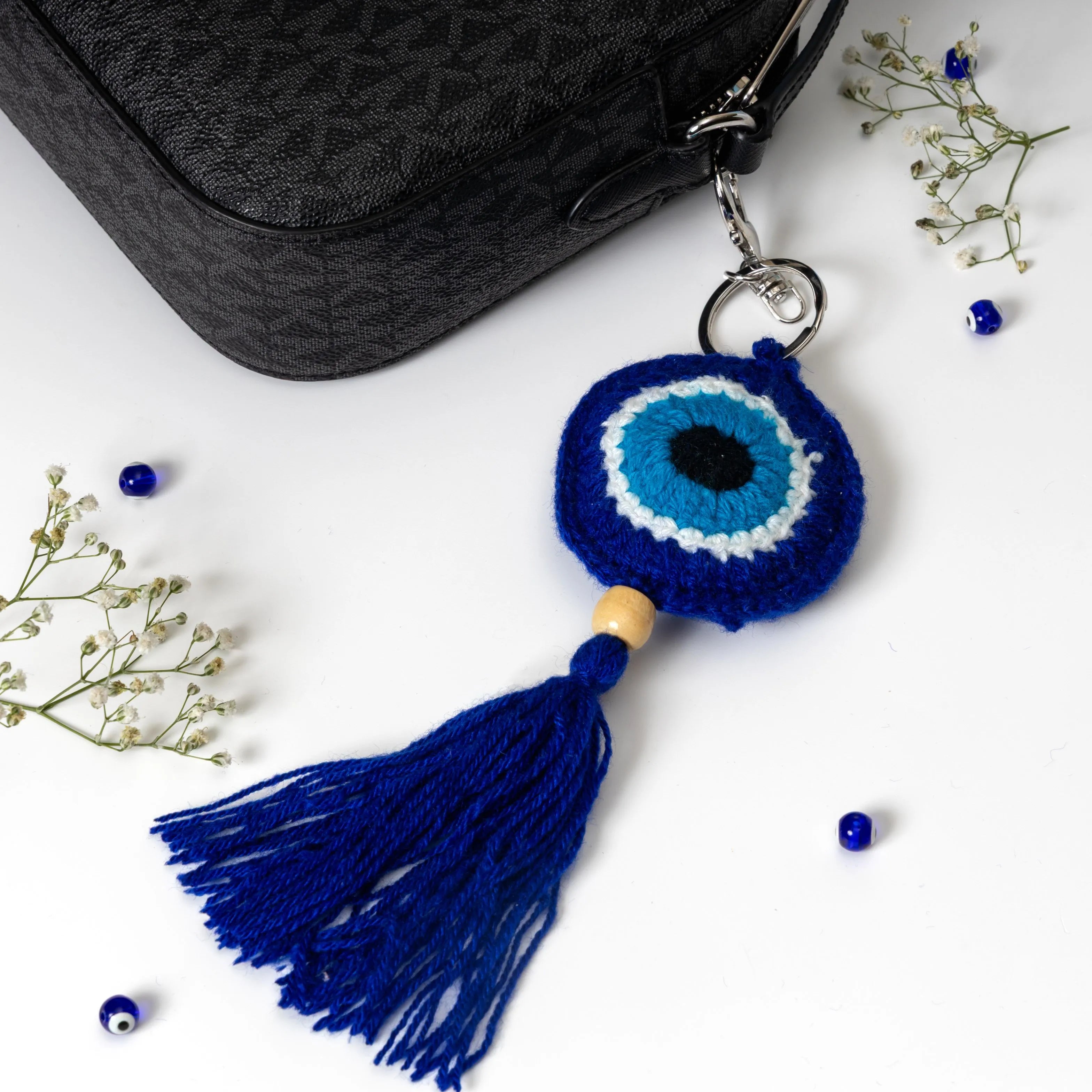 A crocheted evil eye keychain with a large blue tassel, displayed next to a black textured handbag on a white surface with decorative baby's breath and blue beads.