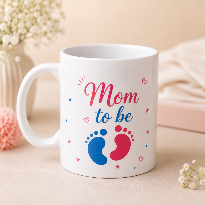 White ceramic "Mom To Be" mug with pink and blue baby footprints, professionally photographed on a soft pastel background for a baby shower gift hamper.