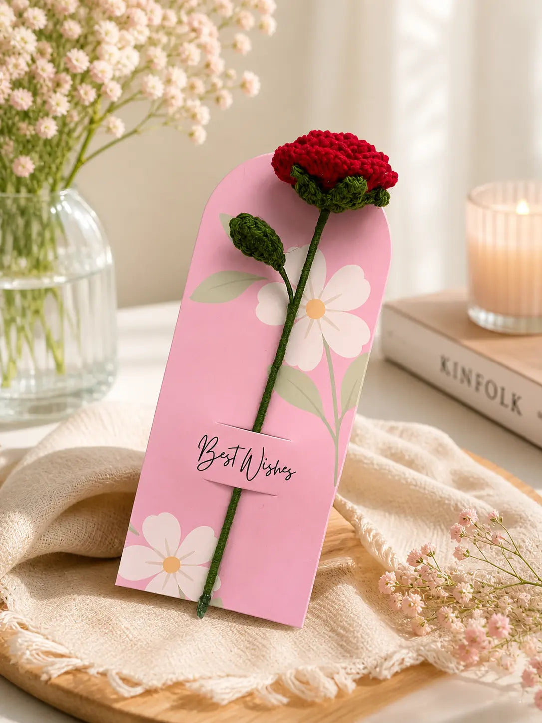 A handmade crocheted flower card with a red carnation and green stem on a pink floral backing, standing on a beige cloth with dried flowers and a candle in the background.