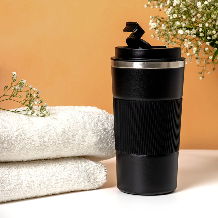Black travel mug with a silver rim on a white surface with white towels and flowers in the background.